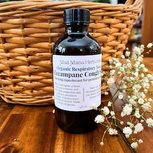 Puede incluir: Una botella de vidrio oscuro de Mad Mama Herbworks Organic Respiratory Tonic Elecampane Cough Syrup. La etiqueta incluye instrucciones y el texto "Warming expectorant for persistent coughs." La botella contiene 118 ml de líquido.