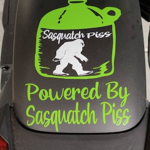 May include: A black and green sticker on a black vehicle fender. The sticker features a green cartoon jug with the text "Sasquatch Piss" and a white cartoon sasquatch walking. Below the jug is the text "Powered By Sasquatch Piss".