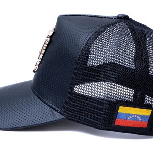 May include: Black leather and mesh trucker hat with a gold-toned embellishment on the front and a Venezuelan flag patch on the back.