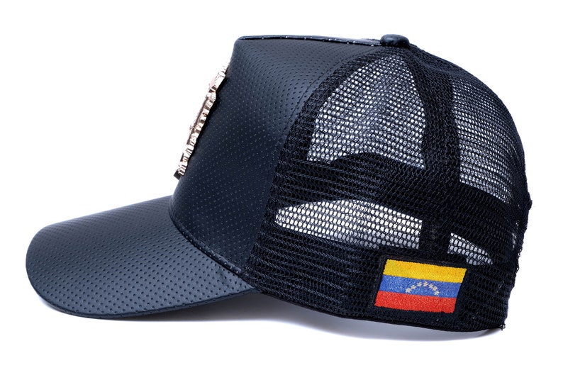 May include: Black leather and mesh trucker hat with a gold-toned embellishment on the front and a Venezuelan flag patch on the back.