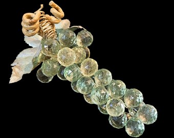 May include: A decorative cluster of grapes with faceted, clear green glass orbs. The grapes are attached to a stem with a gold-colored spiral and fabric leaves. The background is solid black.