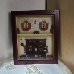 May include: A framed, three-dimensional artwork depicting a floral shop. The shop has a brick facade, a sign that reads "Floral Shop", and a display of flowers. Above the shop are two windows with shutters. The frame is a dark reddish-brown color.