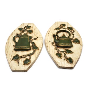 May include: Two decorative wall plaques featuring green teapots and vines on a cream-colored oval background. The teapots have detailed designs and are surrounded by green leaves and stems. The plaques are made of a textured material.