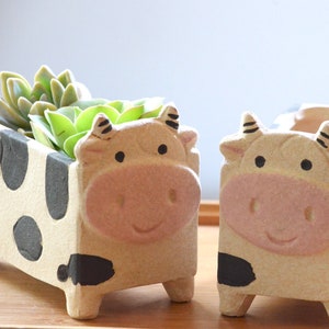 May include: Two ceramic cow-shaped planters. The planters are beige with black spots and feature a cow face design on the front. One planter holds a green succulent plant. The planters are on a wooden surface.