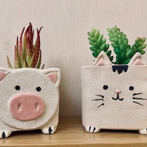 May include: Two ceramic planters shaped like a pig and a cat, both with painted faces. The pig planter has a red and green succulent plant inside, and the cat planter has a green succulent plant inside.