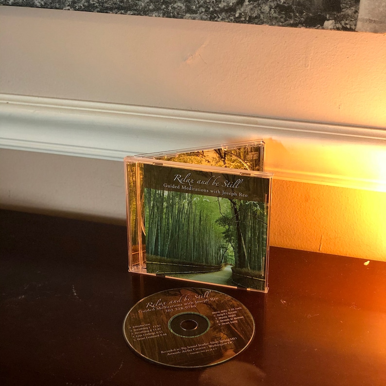 May include: A CD case with a green and white image of a bamboo forest path. The title of the CD is "Relax and be Still" with the subtitle "Guided Meditations with Joseph Men".