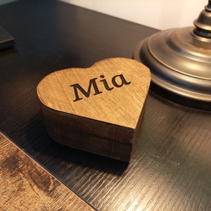 May include: A wooden heart-shaped box with the name "Mia" engraved on the front. The box is sitting on a dark wooden surface.