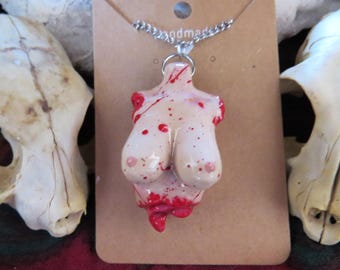 Könnte beinhalten: Eine Halskette mit einem Anhänger in Form eines weiblichen Torsos mit roten Blutflecken und tropfendem Blut. Der Anhänger ist aus weißer Keramik und an einer silbernen Kette befestigt.