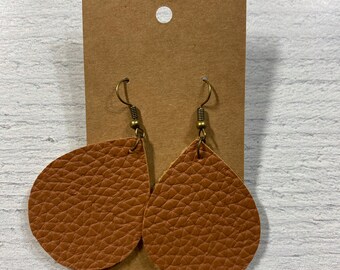 Peut inclure: Une paire de boucles d'oreilles en forme de goutte d'eau de couleur beige, avec une surface texturée. Elles sont suspendues à des crochets de couleur laiton antique et sont présentées sur une carte marron.