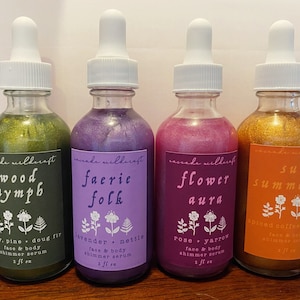 May include: Four glass bottles with dropper tops filled with shimmery liquids. Each bottle has a label with a nature-themed name and ingredients. The labels are: Wood Nymph, Faerie Folk, Flower Aura, and Sun Summon. The bottles are arranged in a row from left to right.
