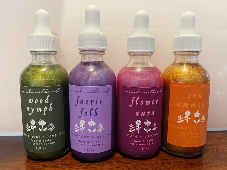 May include: Four glass bottles with dropper tops filled with shimmery liquids. Each bottle has a label with a nature-themed name and ingredients. The labels are: Wood Nymph, Faerie Folk, Flower Aura, and Sun Summon. The bottles are arranged in a row from left to right.