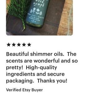 以下が含まれることがあります： 黒いラベルに「wood nymph」と「cedar, pine - doug fir face & body shimmer serum 1 fl oz」と書かれた、小さな透明なガラス瓶。瓶は茶色の表面に緑の葉っぱと一緒に置かれている。