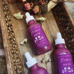 K&ouml;nnte beinhalten: Drei Glasflaschen mit rosa Fl&uuml;ssigkeit und wei&szlig;en Tropfern. Die Flaschen tragen Etiketten mit der Aufschrift "cascade wildcraft flower aura rose + yarrow face & body shimmer serum 2 fl oz".