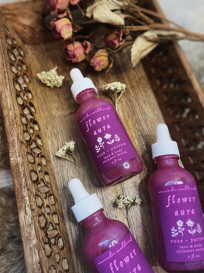 K&ouml;nnte beinhalten: Drei Glasflaschen mit rosa Fl&uuml;ssigkeit und wei&szlig;en Tropfern. Die Flaschen tragen Etiketten mit der Aufschrift "cascade wildcraft flower aura rose + yarrow face & body shimmer serum 2 fl oz".