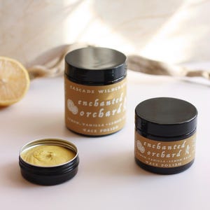 May include: Two black jars of face polish with the label "enchanted orchard" and the text "lemon, vanilla, lemon balm face polish". A small open jar of face polish is in the foreground.