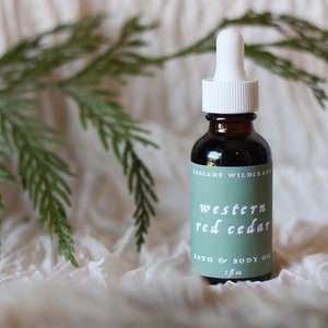May include: A glass bottle with a white dropper top and a green label that reads "CASCADE WILDCRAFT WESTERN RED CEDAR BATH & BODY OIL 1 fl oz".
