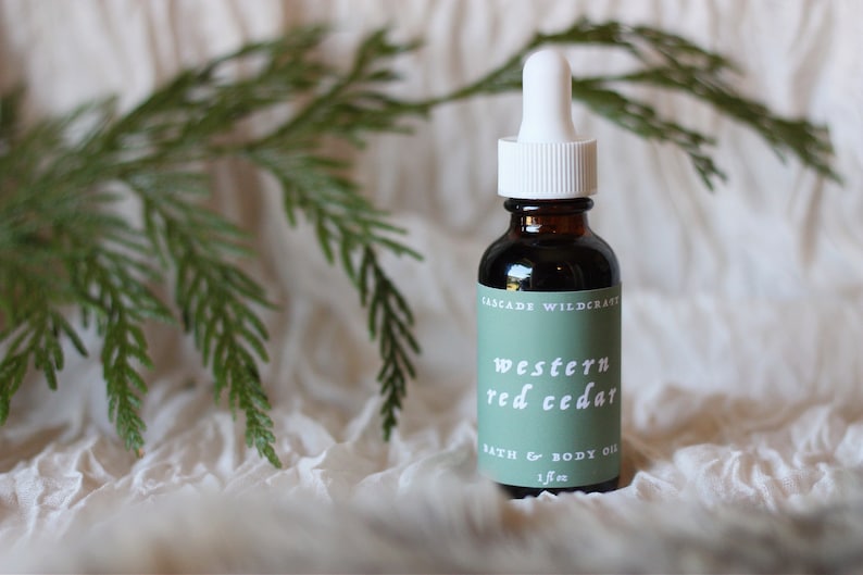 May include: A glass bottle with a white dropper top and a green label that reads "CASCADE WILDCRAFT WESTERN RED CEDAR BATH & BODY OIL 1 fl oz".