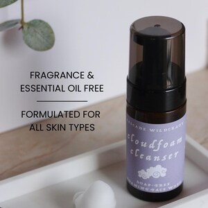 May include: A black and brown bottle of cloudfoam cleanser with a purple label, sitting on a white tray with a white foam and dried plant. The text on the bottle reads "Fragrance & Essential Oil Free" and "Formulated for All Skin Types."