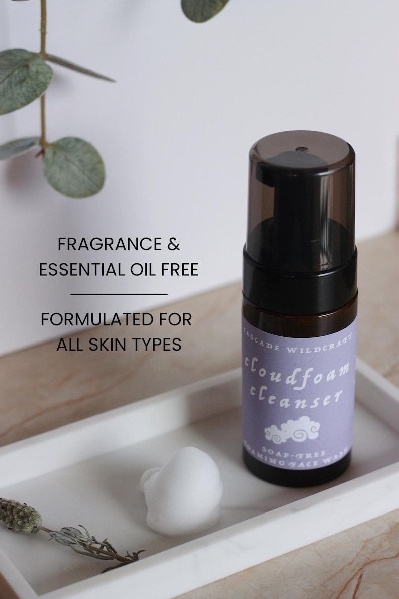 May include: A black and brown bottle of cloudfoam cleanser with a purple label, sitting on a white tray with a white foam and dried plant. The text on the bottle reads "Fragrance & Essential Oil Free" and "Formulated for All Skin Types."