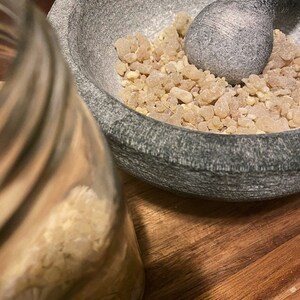 May include: A gray stone mortar and pestle with a pile of light brown resin inside. The mortar and pestle are sitting on a wooden surface.