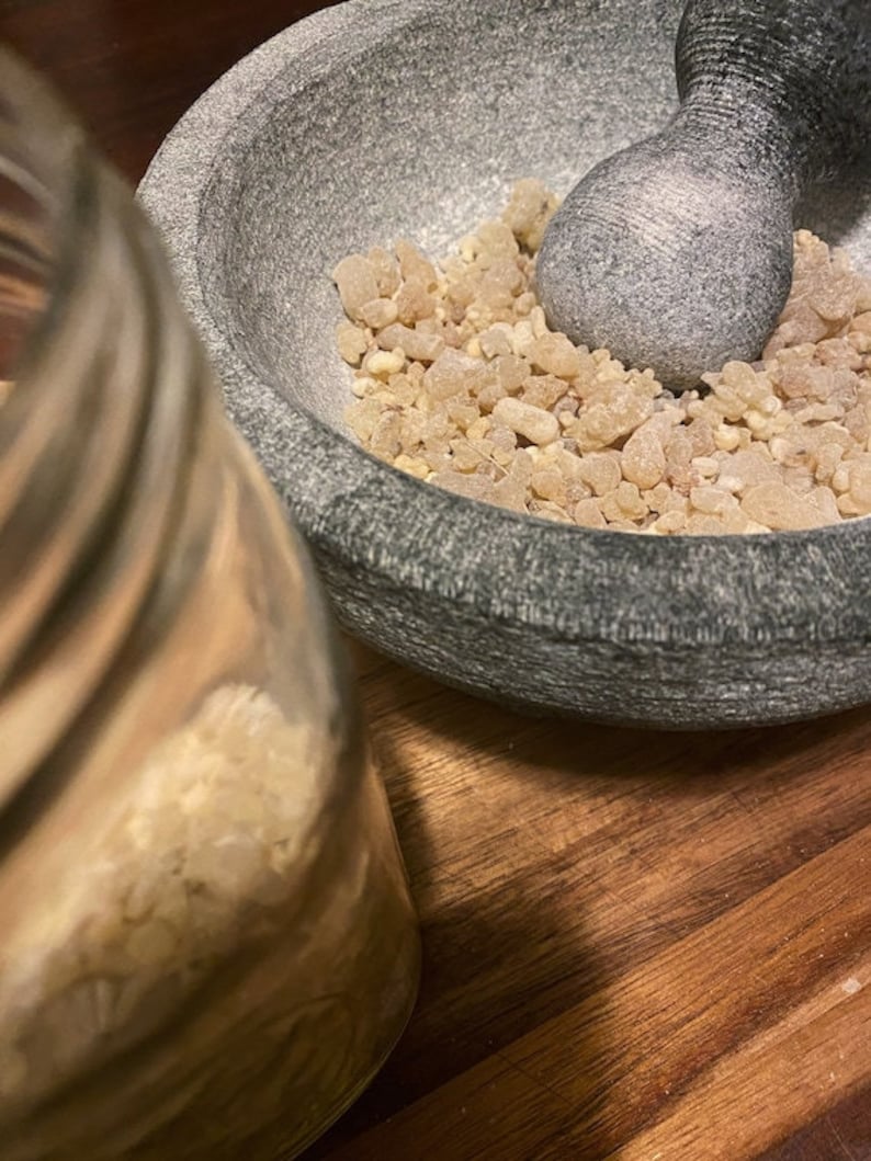 May include: A gray stone mortar and pestle with a pile of light brown resin inside. The mortar and pestle are sitting on a wooden surface.