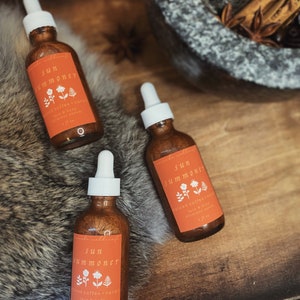May include: Three amber glass bottles with gold shimmery liquid and dropper tops. The bottles are labeled "Sun Summoner" with the text "Spiced Coffee + Cacao Face & Body Shimmer Serum 1 fl oz".