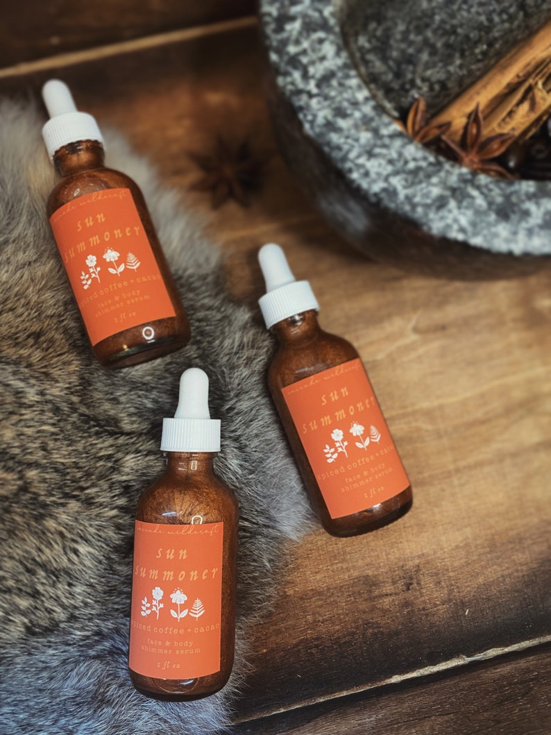 May include: Three amber glass bottles with gold shimmery liquid and dropper tops. The bottles are labeled "Sun Summoner" with the text "Spiced Coffee + Cacao Face & Body Shimmer Serum 1 fl oz".