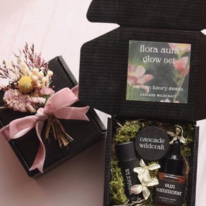 May include: Two black gift boxes, one tied with a pink ribbon and adorned with dried flowers. The other box is open, revealing skincare products, including a bronzing drops bottle, a jar, and a tube. The box also contains a card that reads "flora aura glow set."
