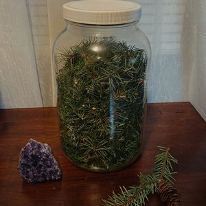 以下が含まれることがあります： 白い蓋が付いた透明なガラス瓶に、新鮮な緑色の松の葉が詰められています。紫色のアメジストの結晶と松の枝に松ぼっくりが、木製の表面の上に置かれています。
