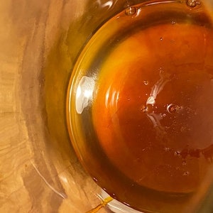 May include: A close-up view of a glass jar filled with a thick, amber-colored liquid. The liquid has a slightly cloudy appearance and is illuminated by a bright light source.