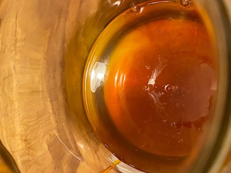 May include: A close-up view of a glass jar filled with a thick, amber-colored liquid. The liquid has a slightly cloudy appearance and is illuminated by a bright light source.
