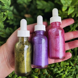 K&ouml;nnte beinhalten: Drei Glasflaschen mit Tropfern, gef&uuml;llt mit glitzernder Fl&uuml;ssigkeit. Die Flaschen sind gold, lila und rosa.