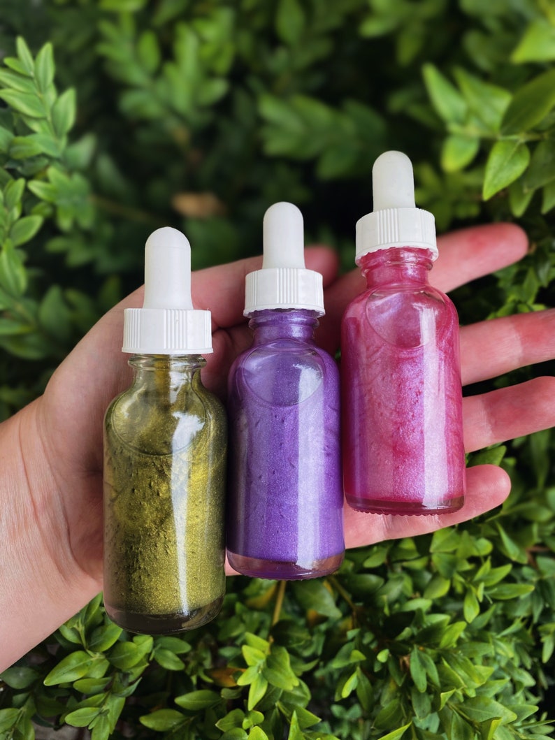 K&ouml;nnte beinhalten: Drei Glasflaschen mit Tropfern, gef&uuml;llt mit glitzernder Fl&uuml;ssigkeit. Die Flaschen sind gold, lila und rosa.