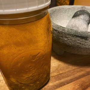 May include: A clear glass jar with a white lid filled with a yellow liquid and small white pieces. The jar is sitting on a wooden surface next to a gray stone mortar and pestle.
