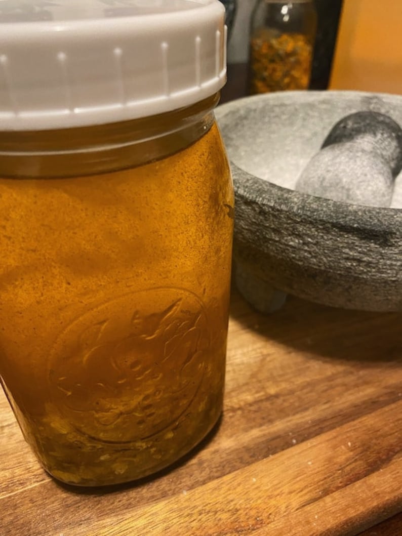 May include: A clear glass jar with a white lid filled with a yellow liquid and small white pieces. The jar is sitting on a wooden surface next to a gray stone mortar and pestle.