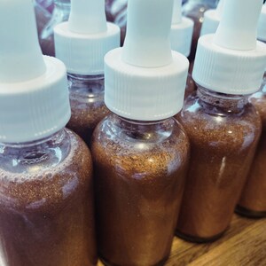 May include: A group of small glass bottles with white dropper tops filled with a shimmering brown liquid. The bottles are arranged in a row on a wooden surface.