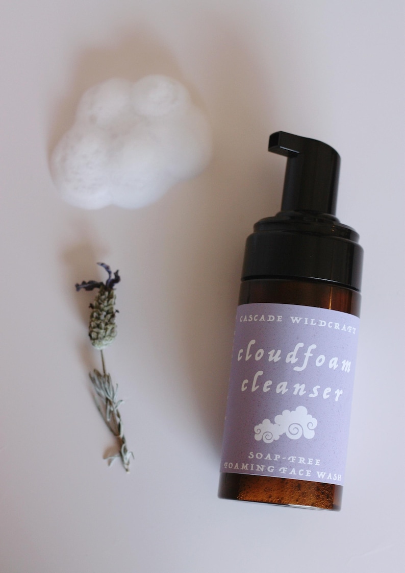 May include: A brown glass bottle of Cascade Wildcraft cloudfoam cleanser with a black pump top. The label is lavender with white text and a cloud graphic. A small cloud of white foam and a sprig of dried lavender are also present.