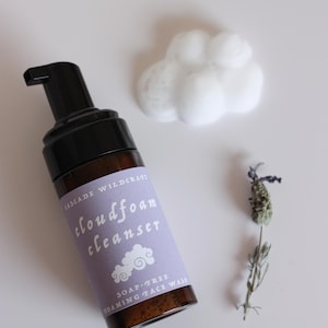 May include: A brown glass bottle of Cloudfoam Cleanser with a black pump, a cloud of white foam, and a sprig of lavender. The label is light purple with white text and a cloud graphic. The product is a soap-free foaming face wash.
