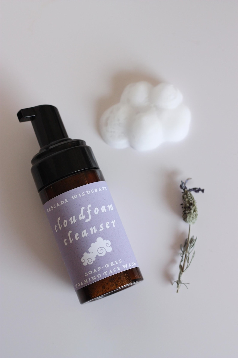 May include: A brown glass bottle of Cloudfoam Cleanser with a black pump, a cloud of white foam, and a sprig of lavender. The label is light purple with white text and a cloud graphic. The product is a soap-free foaming face wash.