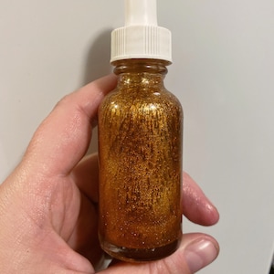 May include: A glass bottle with a gold liquid and glitter inside. The bottle has a white dropper top.