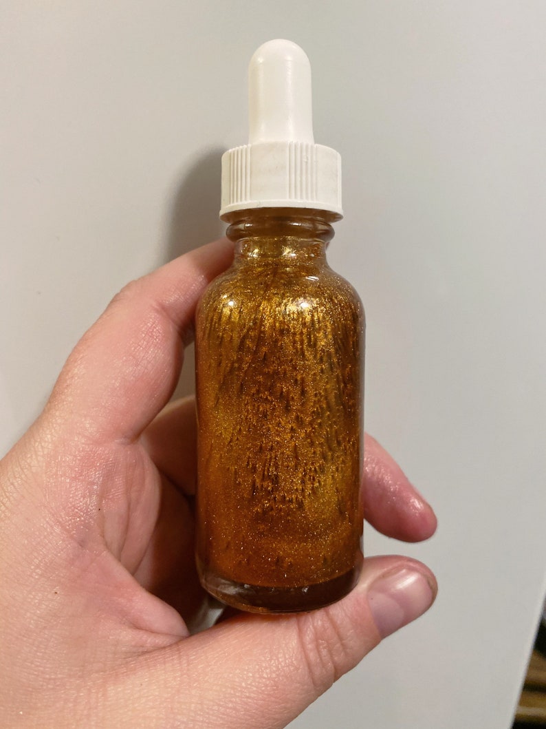 May include: A glass bottle with a gold liquid and glitter inside. The bottle has a white dropper top.