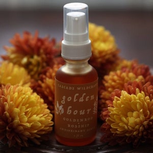 May include: A glass bottle with a white pump top and a label that reads "Golden Hour Goldenrod Rosehip Antioxidant Serum 1 fl oz". The bottle is surrounded by yellow and orange flowers.