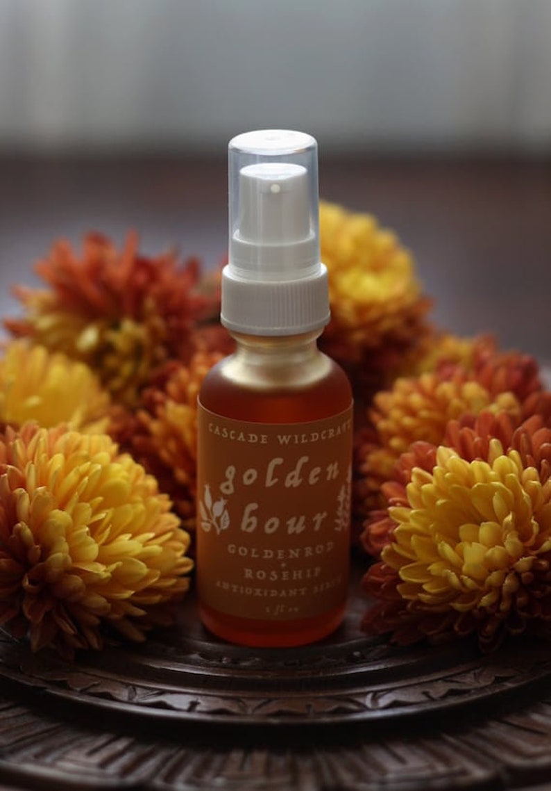 May include: A glass bottle with a white pump top and a label that reads "Golden Hour Goldenrod Rosehip Antioxidant Serum 1 fl oz". The bottle is surrounded by yellow and orange flowers.
