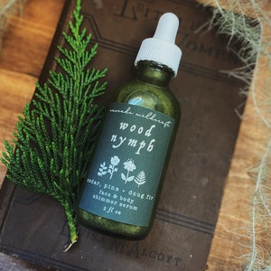 以下が含まれることがあります： ゴールドの輝きを帯びた緑色のガラス瓶で、「wood nymph」と「cedar, pine + doug fir face & body shimmer serum 2 fl oz」のラベルが貼られています。