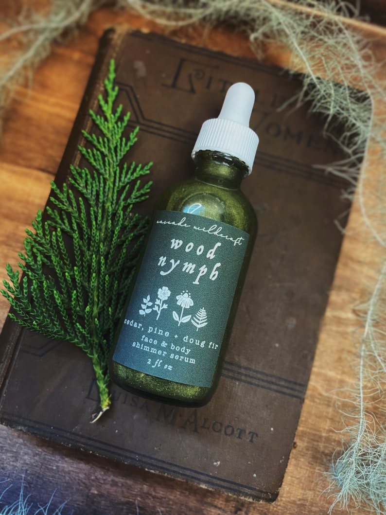 以下が含まれることがあります： ゴールドの輝きを帯びた緑色のガラス瓶で、「wood nymph」と「cedar, pine + doug fir face & body shimmer serum 2 fl oz」のラベルが貼られています。
