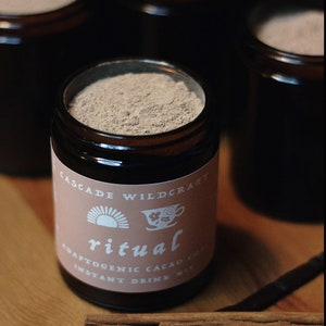 May include: A dark brown glass jar filled with a light brown powder, labeled "ritual" with the text "Adaptogenic Cacao Chai Instant Drink Mix." Cinnamon sticks and a vanilla bean are in the foreground. Other jars are in the background.