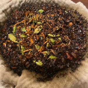May include: A white cloth bag filled with dark coffee grounds, cardamom pods, and other spices. The bag is being used to filter the coffee.
