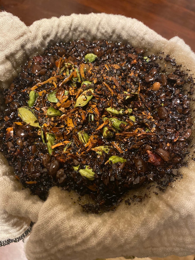 May include: A white cloth bag filled with dark coffee grounds, cardamom pods, and other spices. The bag is being used to filter the coffee.