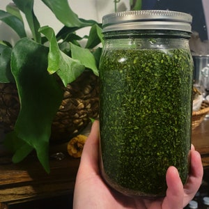 May include: A glass jar filled with a dark green, finely chopped herb mixture. The jar has a silver lid and is being held by a hand.