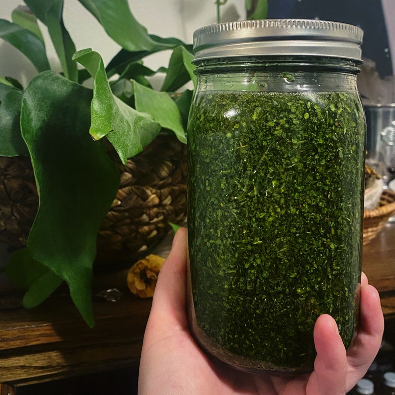 May include: A glass jar filled with a dark green, finely chopped herb mixture. The jar has a silver lid and is being held by a hand.